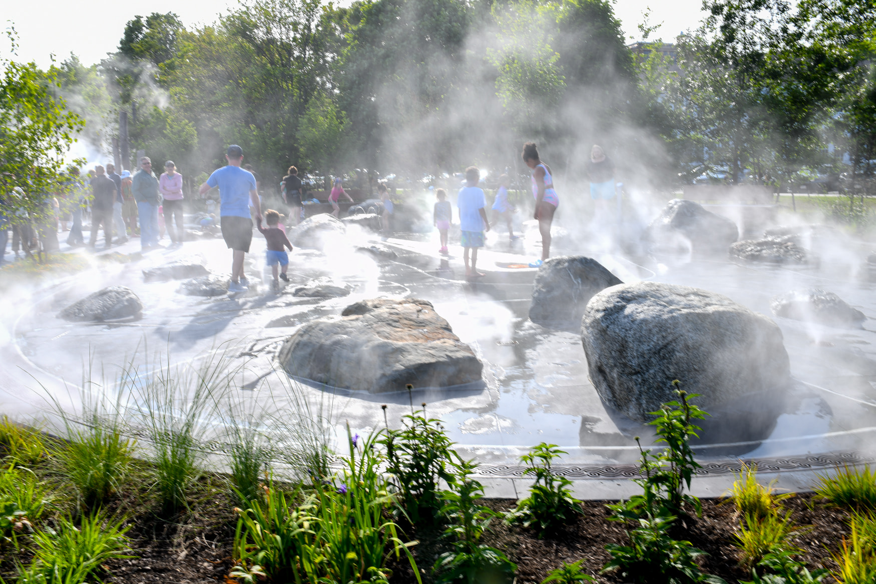 Children and families enjoying a splash park at the Pattern District in Newton Upper Falls, MA, featuring interactive mist and water play areas.